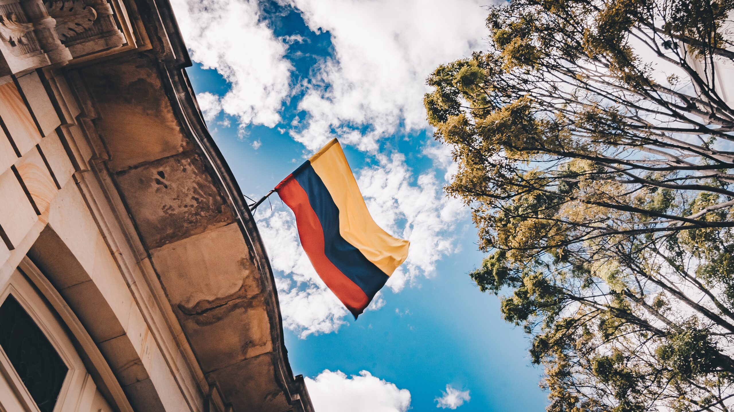 Vista do céu azul com algumas nuvens, à direita uma árvore e à esqueda uma casa com uma bandeira da Colombia.