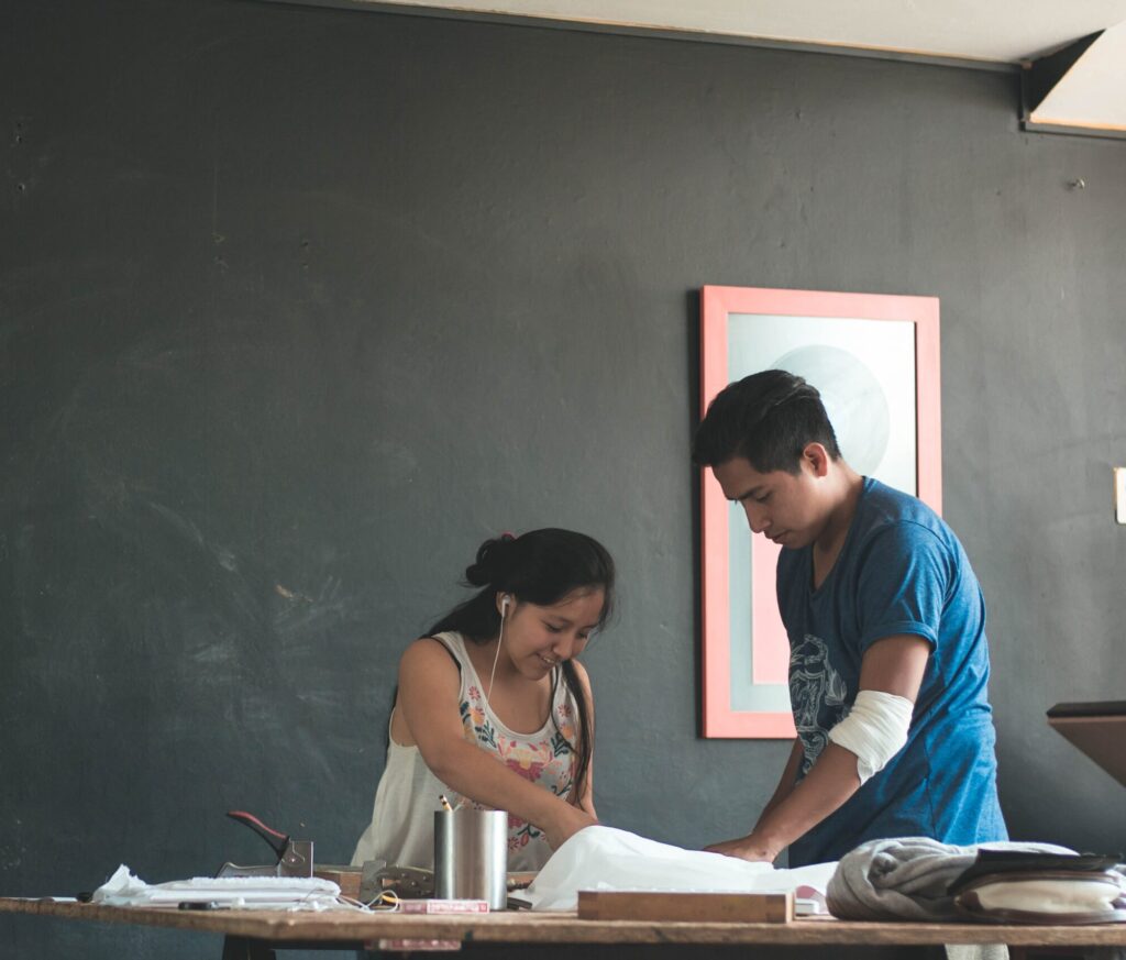 Em uma sala de aula com um quadro negro, estão uma aluna e um professor conversando sobre um exercício na mesa. O professor está com o cotovelo enfaixado.