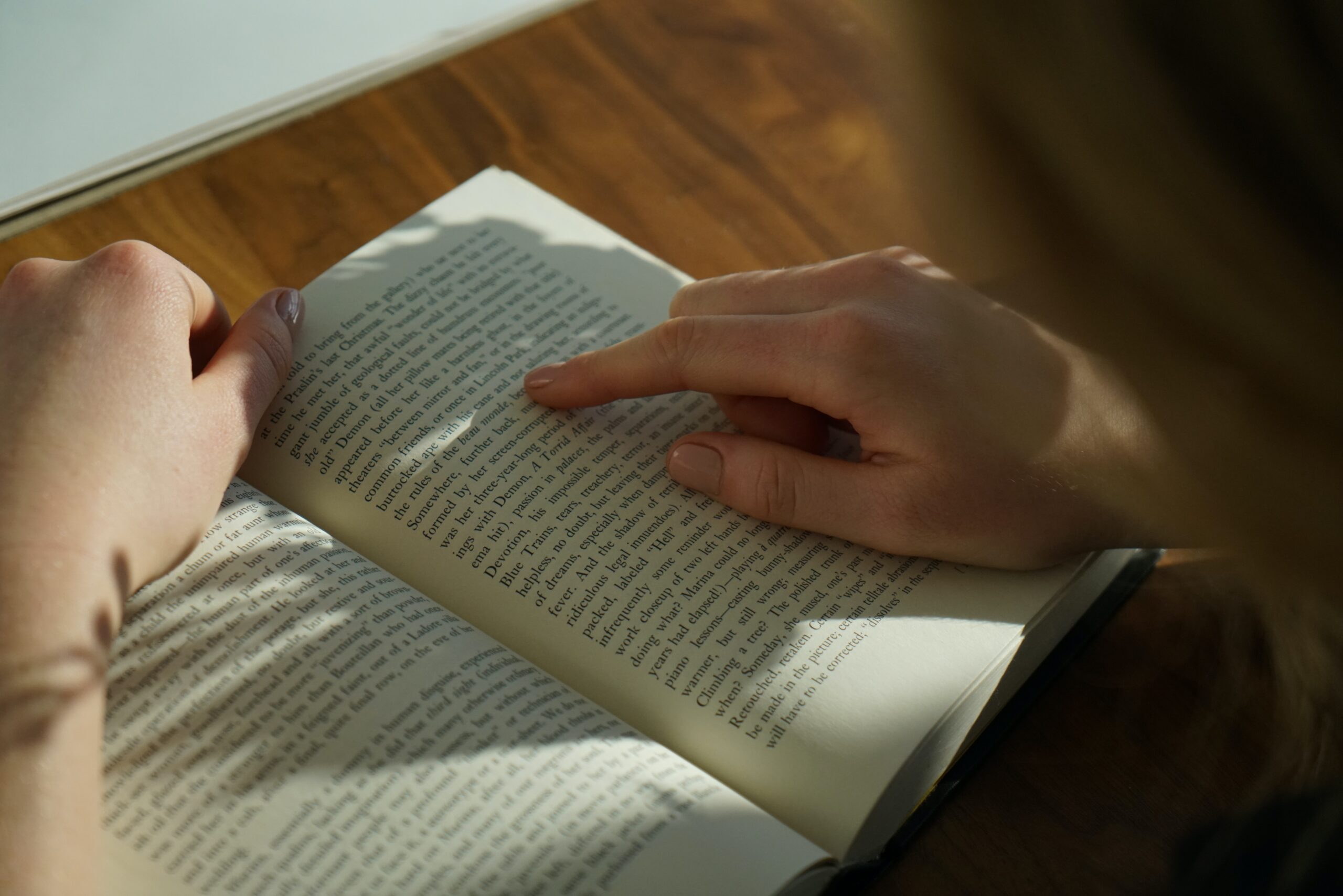 Um livro aberto, com a sombra da cabeça da pessoa lendo. Estão visíveis as maõs apenas, e a uma parte da silhueta da cabeça. A pessoa está acompanhando a leitura com o dedo.