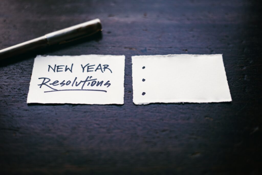 Uma caneta e dois pedaços de papel, o primeiro escrito "New Year Resolutions, e o segundo com 3 pontos para as resoluções serem escritas.